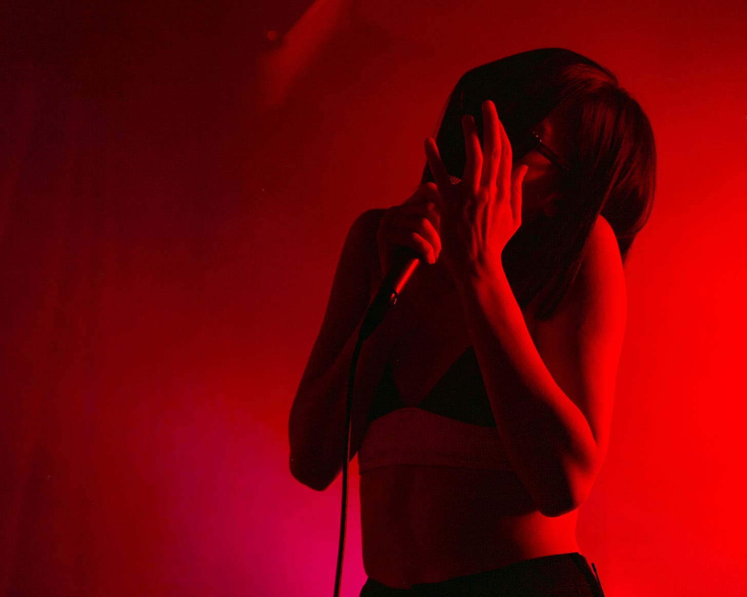 a slim framed human with soft curves sings into a microphone, the photo is red and black due to intensely pigmented red lights