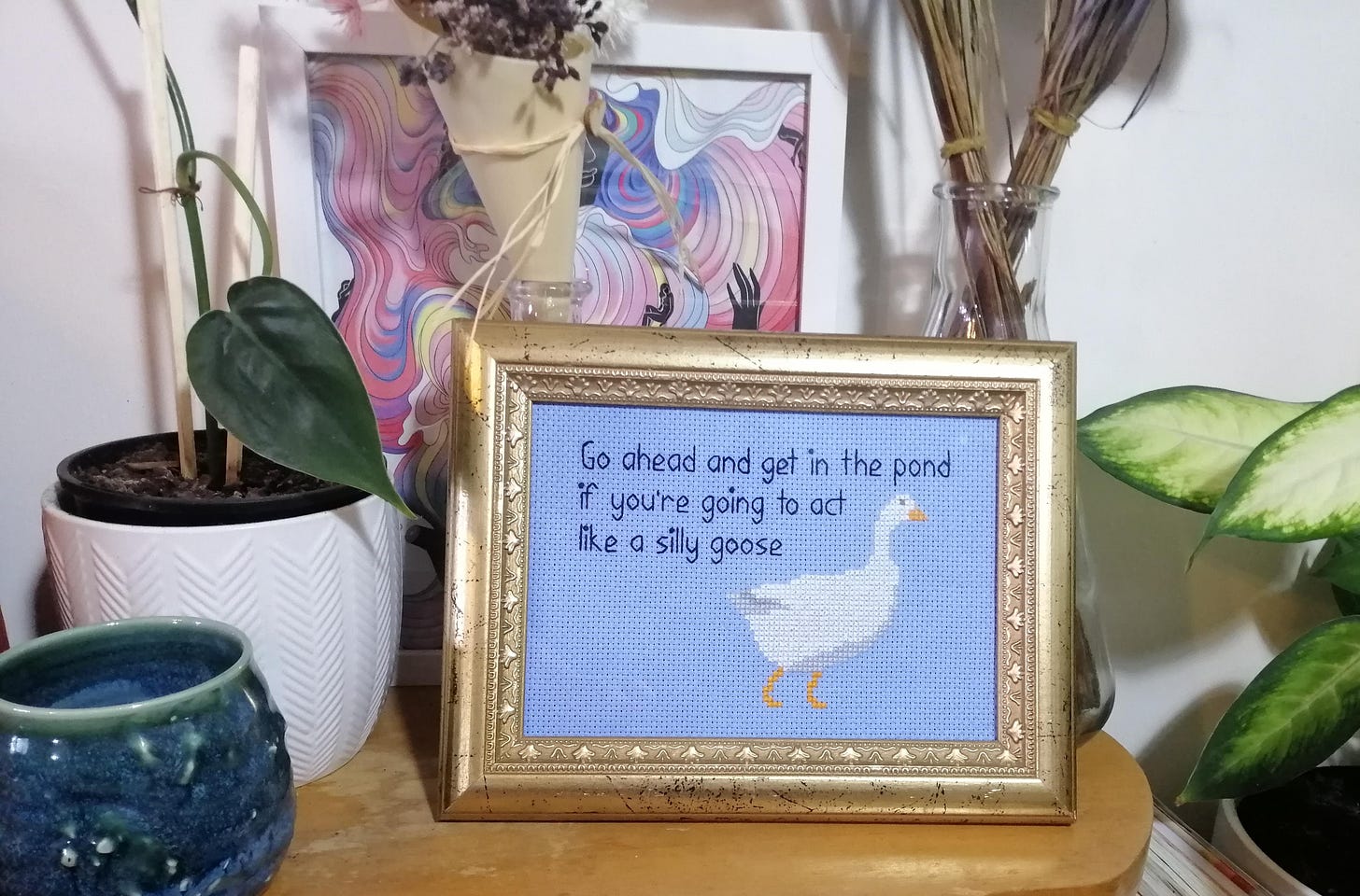 Cross stitch in a gold frame on a shelf with plants and stuff. The stitch is on blue and has a picture of a goose with the text "Go ahead and get in the pond if you're going to act like a silly goose"