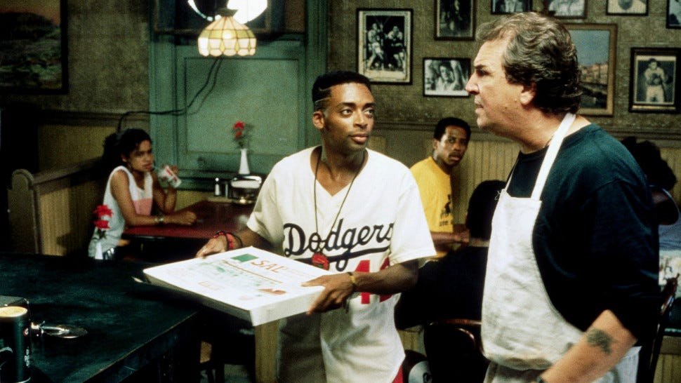 This image shows two of Do the Right Thing's protagonists: On the left, Mookie (played by Spike Lee)--who holds a pizza box in his hands and wears a reproduction of Jackie Robinson's Number 42 Brooklyn Dodgers' jersey--and, on the right, Sal Frangione (played by Danny Aiello), who wears an apron while speaking to Mookie. Three unnamed customers sit in the background.