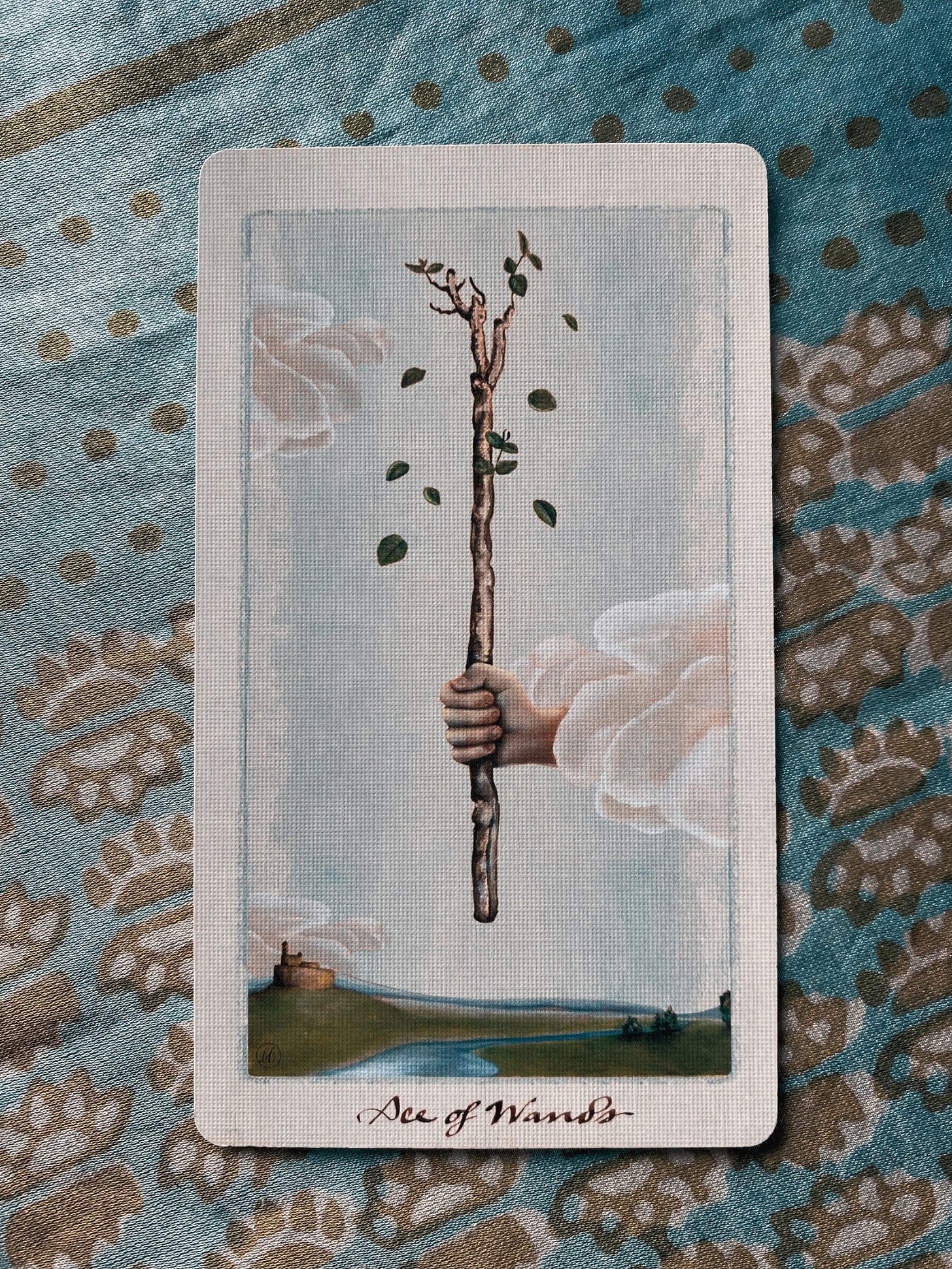 a photo of the ace of wands from the pagan otherworlds tarot laying atop a muted teal silk cloth. the cloth has golden prints of circles and flower-like suns and a golden lined border. the ace of wands card shows a branch upright, held in a firm pale hand that extends from a cloud in a light blue sky. green leaves fall gently from the buds at the top of the branch. below the branch is a river running through a green country, trees in the foreground and one side, and a castle on a hill in the background of the other side. behind the scene, the river flows into the dark blue shadow of far distant mountains.