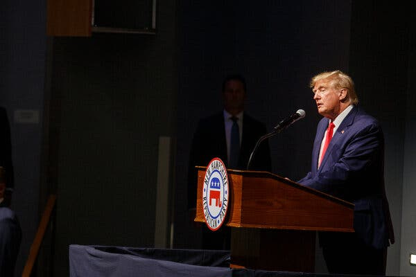 Former President Donald Trump, wearing a blue suit and a red tie, speaks behind a wooden lectern during a recent campaign stop in New Hampshire.