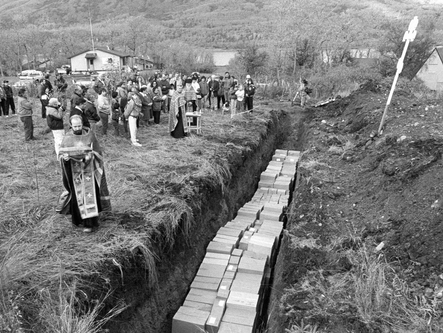 A black and white photo of an open gravesite with dozens of boxes. People stand next to the ground and are gathering for a ceremony.