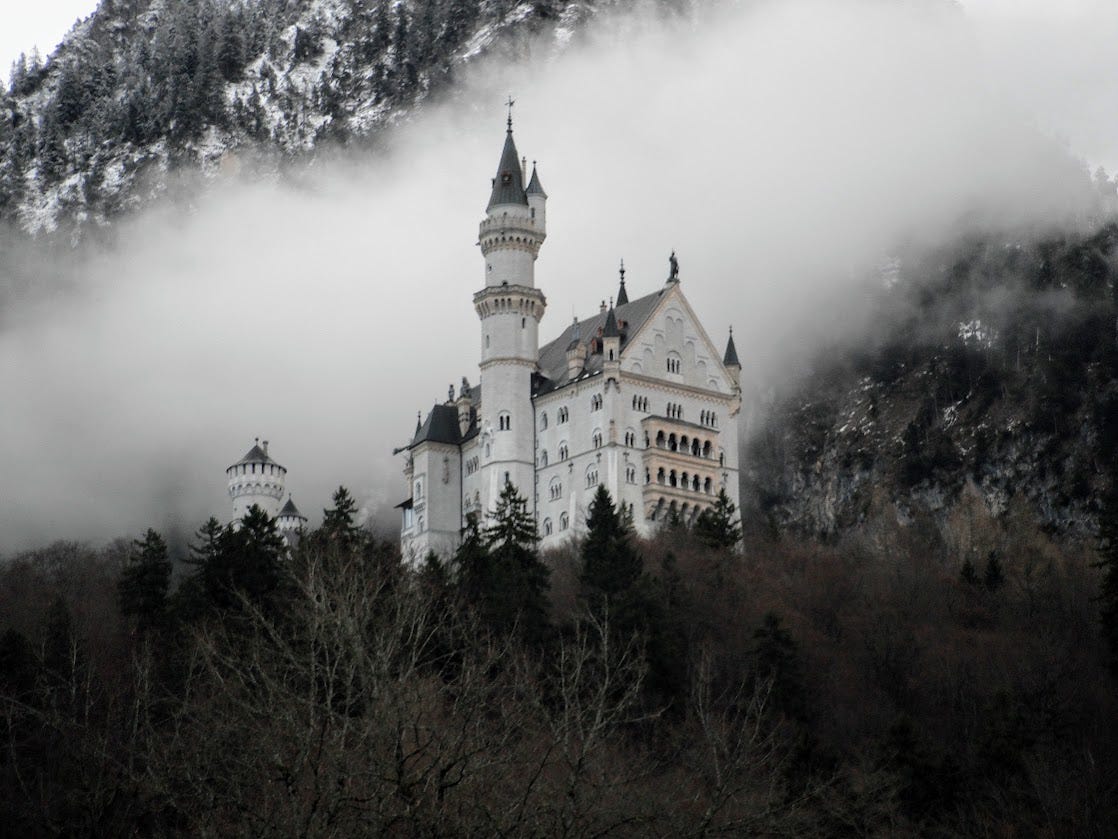 Neuschwanstein, the iconic fairy tale castle in Germany, sits high on a hill, surrounded by clouds and backed by a tree-covered mountain behind.
