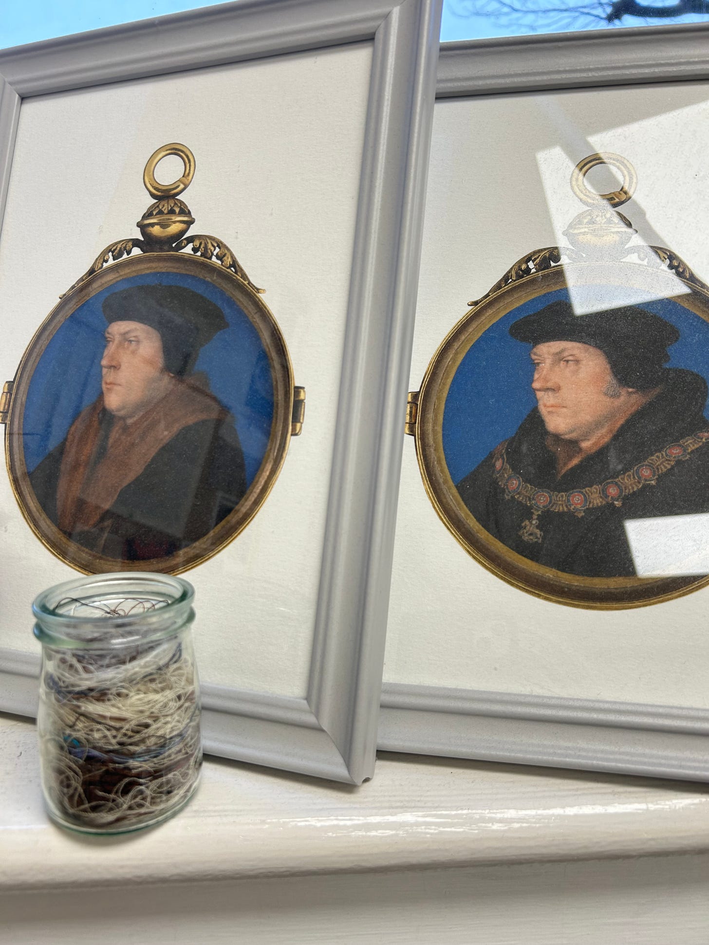 Two portraits of Thomas Cromwell in grey frames, facing a jar of threads in random colours and lengths