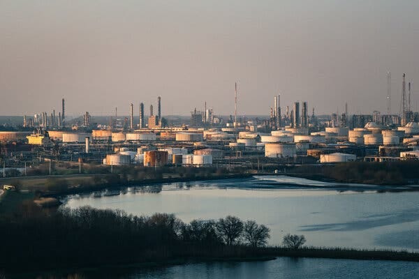 An Exxon Mobil refinery with tanks, smoke stacks and towers in the background. A small body of water is in the foreground.
