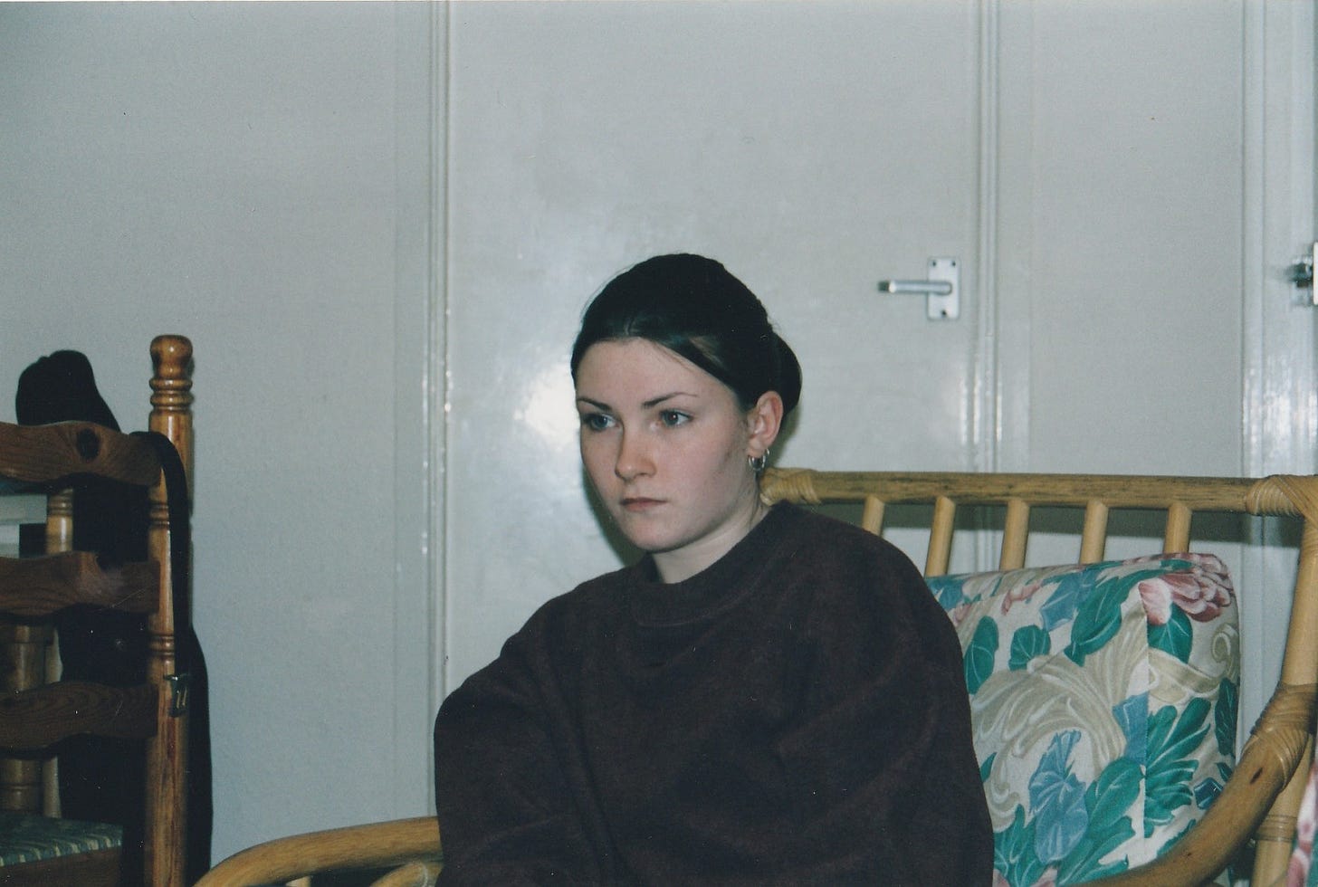 A girl is captured looking into the middle distance. She sits on a wicker chair in a white room