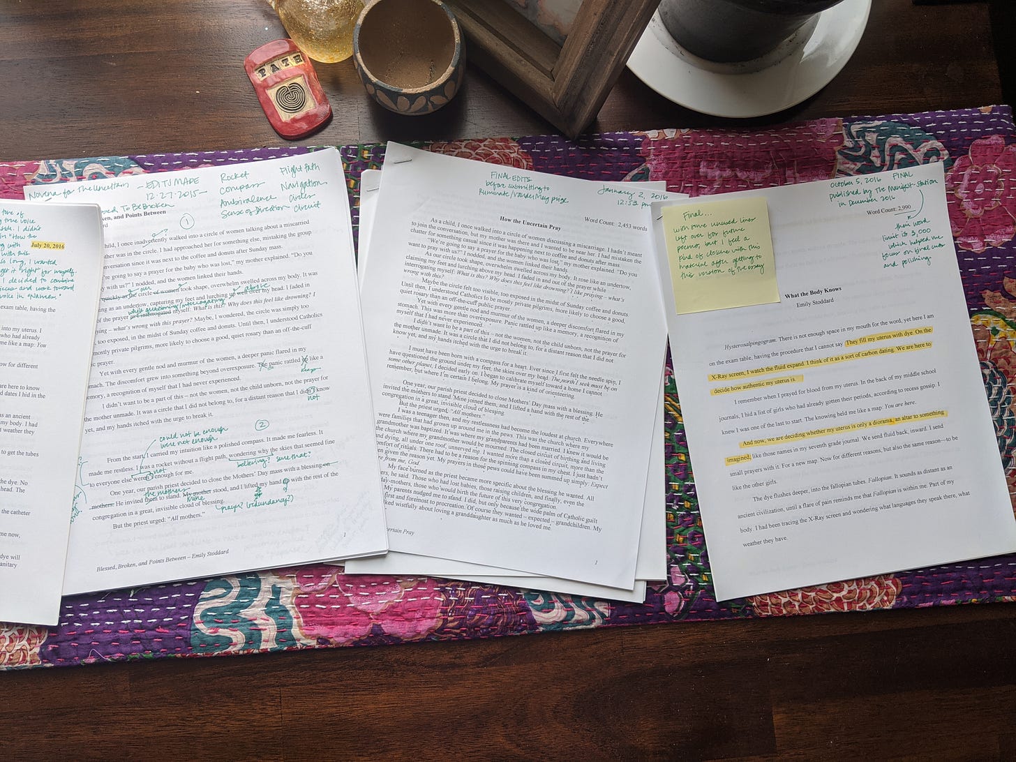 A series of small paper piles spread across a table, on top of a colorful purple runner. The drafts have highlighted parts and marks in green ink, where I've noted changes or themes that I have questions about.