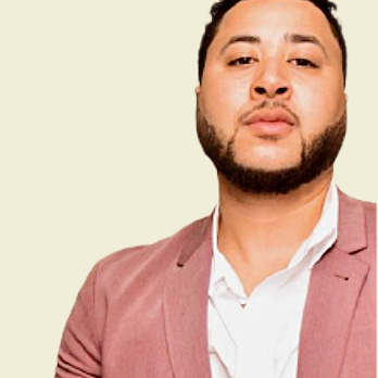 Achieving Goals with Perseverance: Pharaoh Kelley on Health, Real Estate, and Resilience. A man with short curly hair and a beard is wearing a pink blazer over a white collared shirt. He is looking directly at the camera with a neutral expression, embodying resilience. The background is a plain, light beige color.