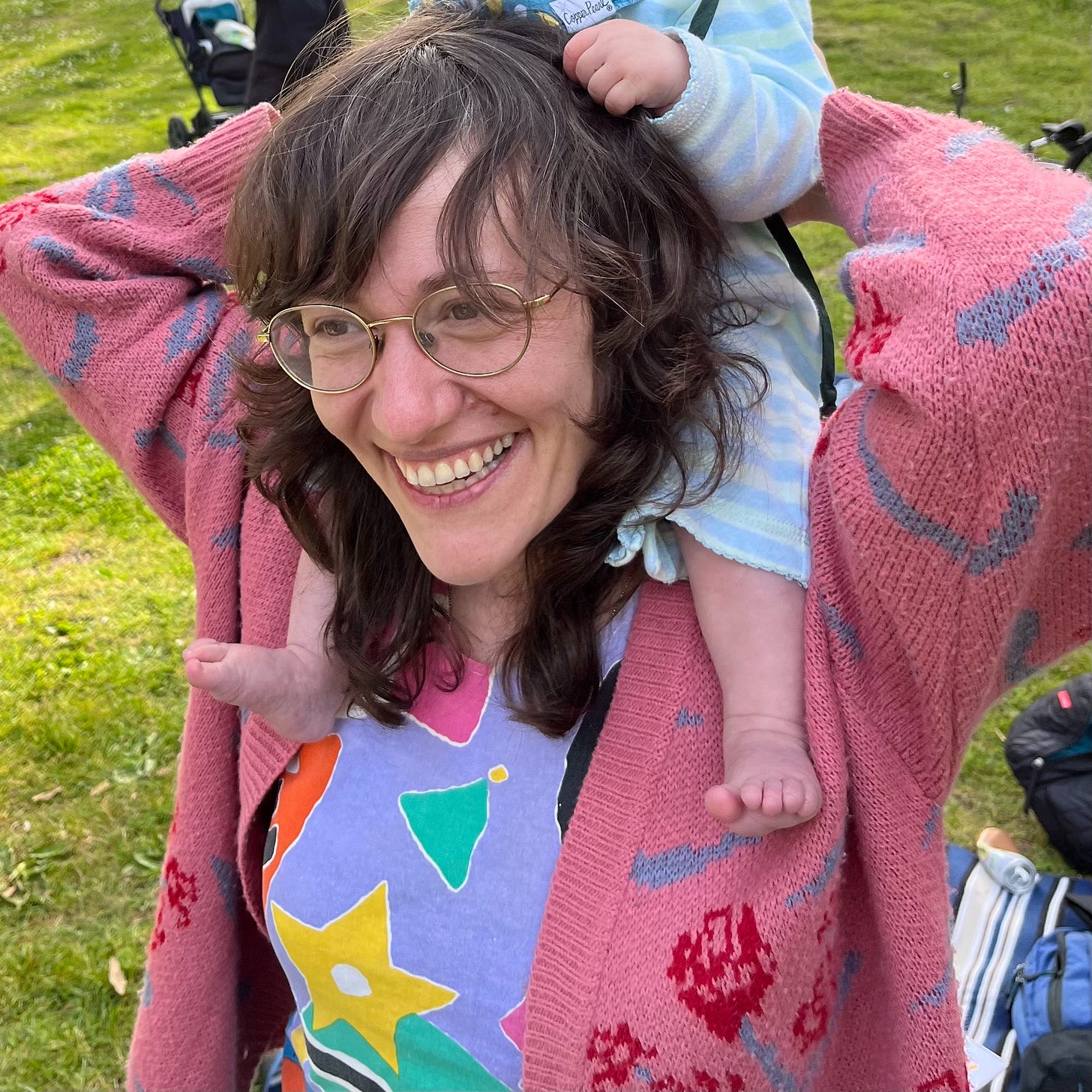 Amy is wearing a purple shirt with colorful shapes and a pink cardigan with roses on it. She's holding her baby on her shoulders and her baby is holding onto her hair like the reins of a horse. Her baby's cute little feet are bare.