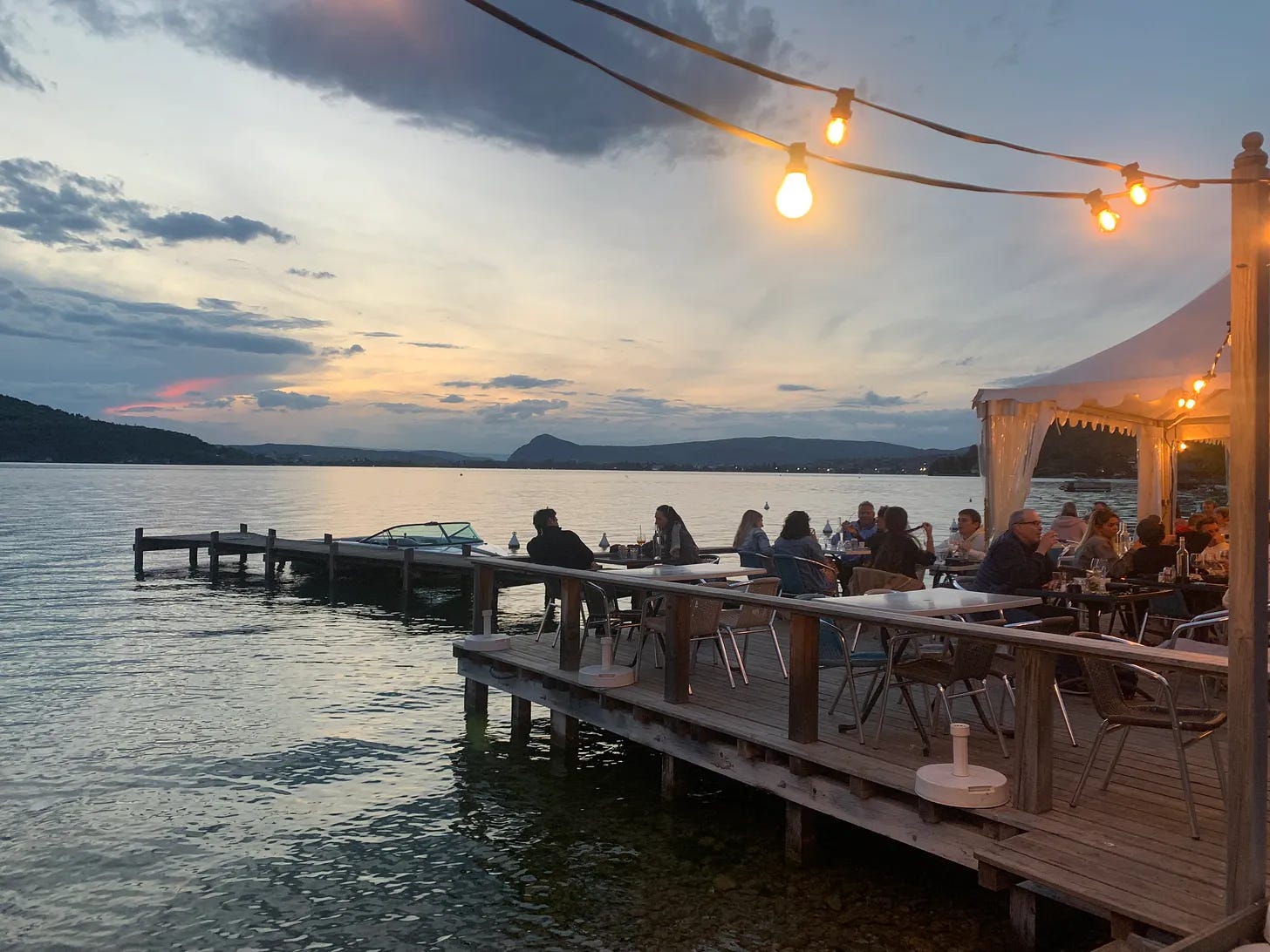 Evening dining at Le Pecheur Restaurant on Lake Annecy in Veyrier-du-Lac, with patrons enjoying meals on a wooden deck over the water, illuminated by string lights, against a backdrop of a picturesque sunset and calm lake.