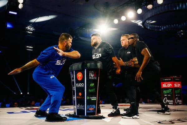 Four men stand on a brightly lit stage. One man dressed in blue faces three others as he swings his arm back in preparation for delivering a slap.