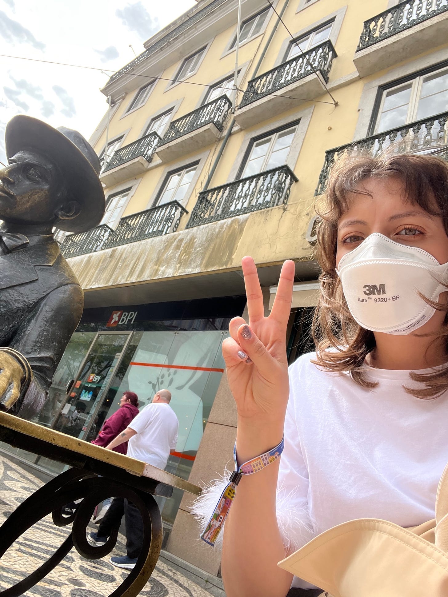 Eu fazendo sinal de paz e amor com os dedos, usando máscara Aura, sentada a uns 3 passos de distância da estátua do Fernando Pessoa, feita em bronze, nas ruas de Lisboa