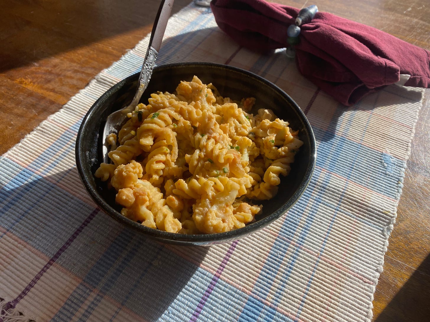 A ceramic bowl of chickpea pasta sitting on a woven placemat next to a red napkin on a sunny table.