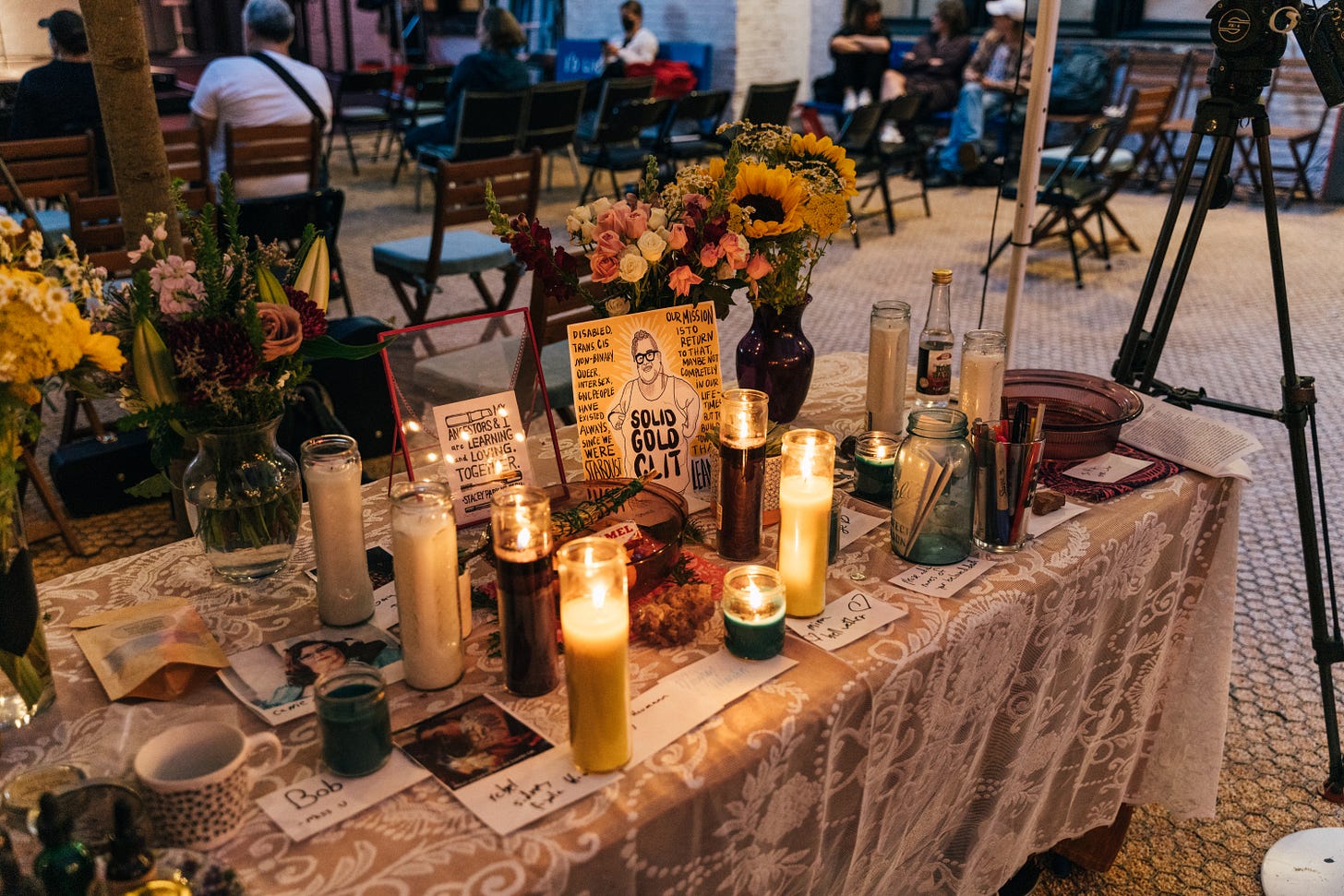 photo of a table altar covered with a white lace cloth, roses and sunflowers, yellow and green 7 day candles, images of disabled loved ones and white pieces of paper with their names on them, in front of an area where people are seated with their back to the viewer, it's twilight.