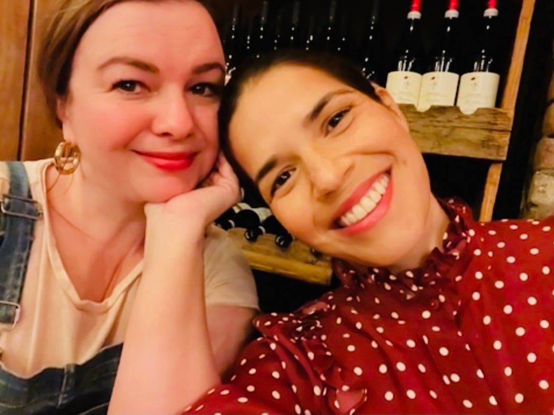 A selfie of Amber Tamblyn and America Ferrera taken from the neck up. America tilts her head to touch Amber's. Both smile.