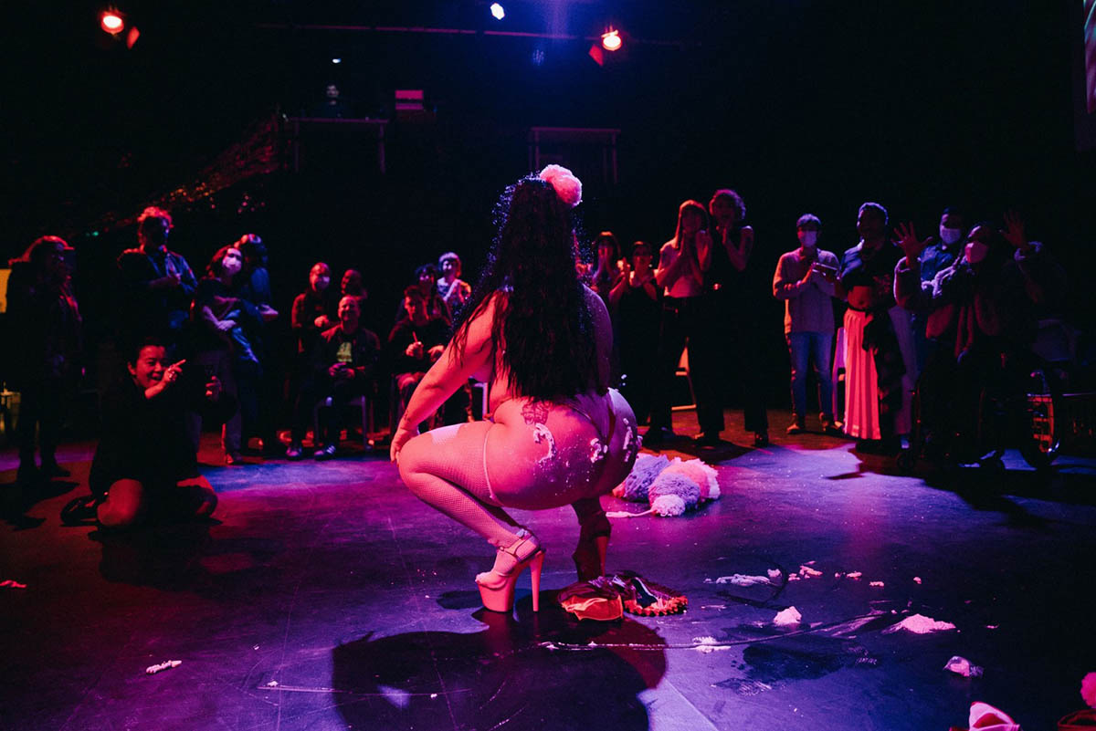 A femme performer wearing a thong and pleaser high heels is bathed in pink and purple light in a dark venue. It looks like she’s just done a cake sit because there is frosting or whipped cream on her ass. A shadowed crowd is living for it, including several manual wheelchair users.