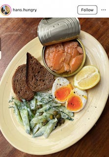 A plate of food from instagram. A white plate, with two small brown toast triangles, a boiled egg halved, a wedge of lemon, an open tin of what looks like chunks of trout or salmon, and a dollop of cucumber salad with capers and mayo.