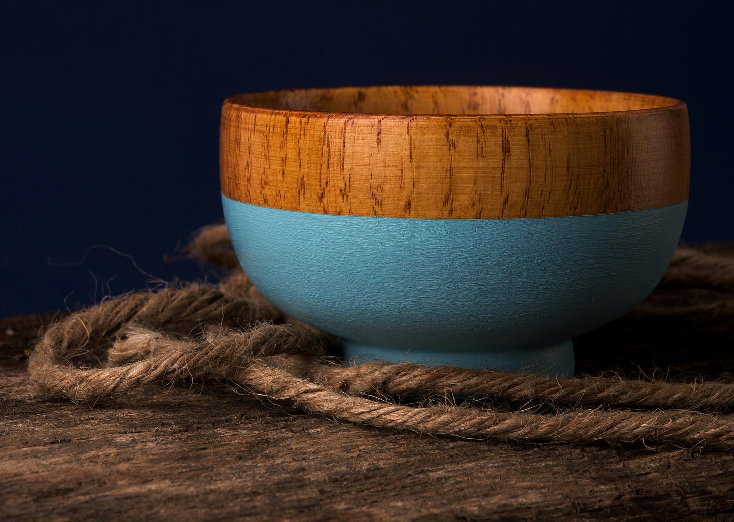 A hand-crafted wooden bowl sitting on a dark wood table, surrounded by rope. The bottom of the bowl is painted a soft blue. The backdrop is like a dark night sky with no stars.