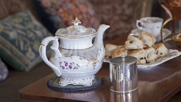An ornate teapot set on a low table with a plate of scones behind