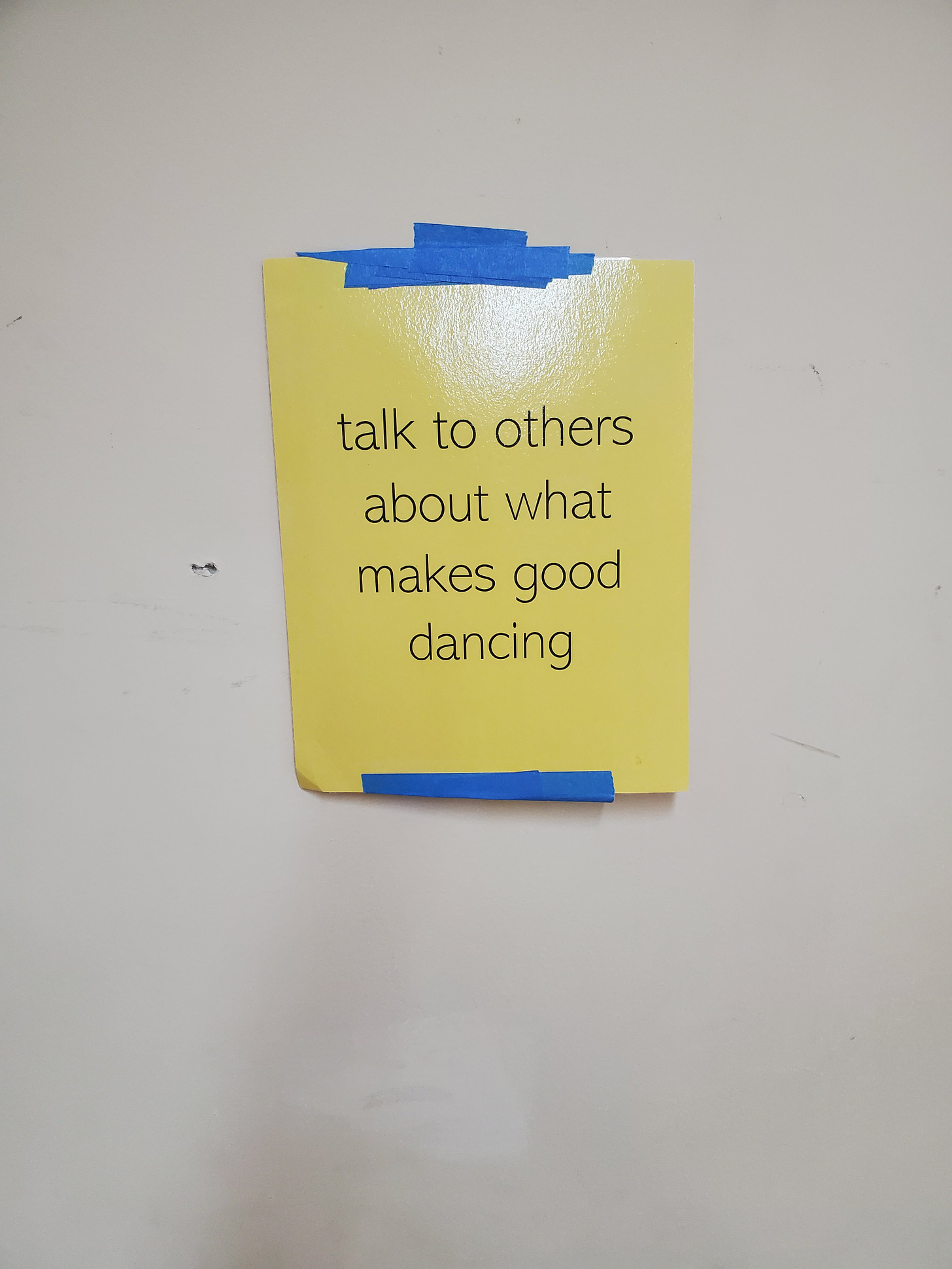 A yellow sign affixed to a white wall with blue painter's tape reads "talk to others about what makes good dancing".