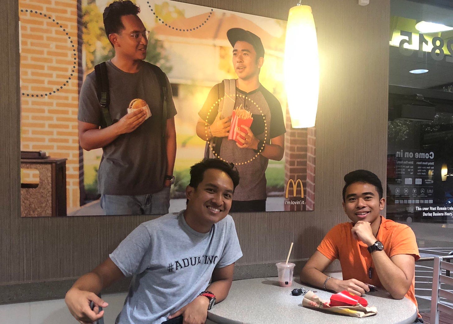 A poster of two men with McDonalds food. Sitting in front of the poster are the same two men in McDonalds