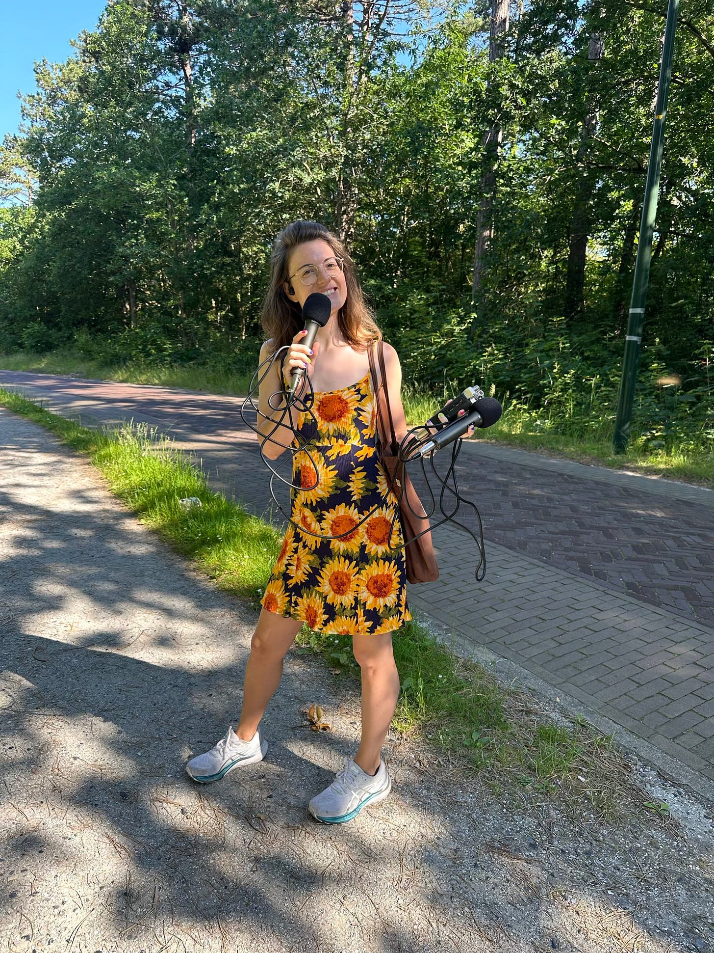 Foto van Nydia, een witte vrouw van in de 30 met lang bruin haar. Ze draagt een bril en heeft een jurkje met geschilderde zonnebloemen er op aan. Ze staat op een pad op straat en heeft een microfoon en een recorder in haar hand, waar ze in praat