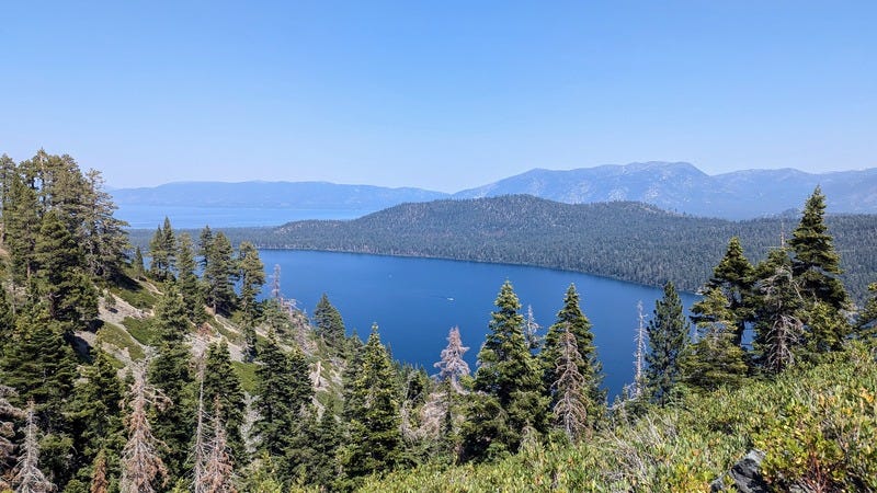 Looking down onto a huge lake from a forested mountainside, sky and mountains in the distance