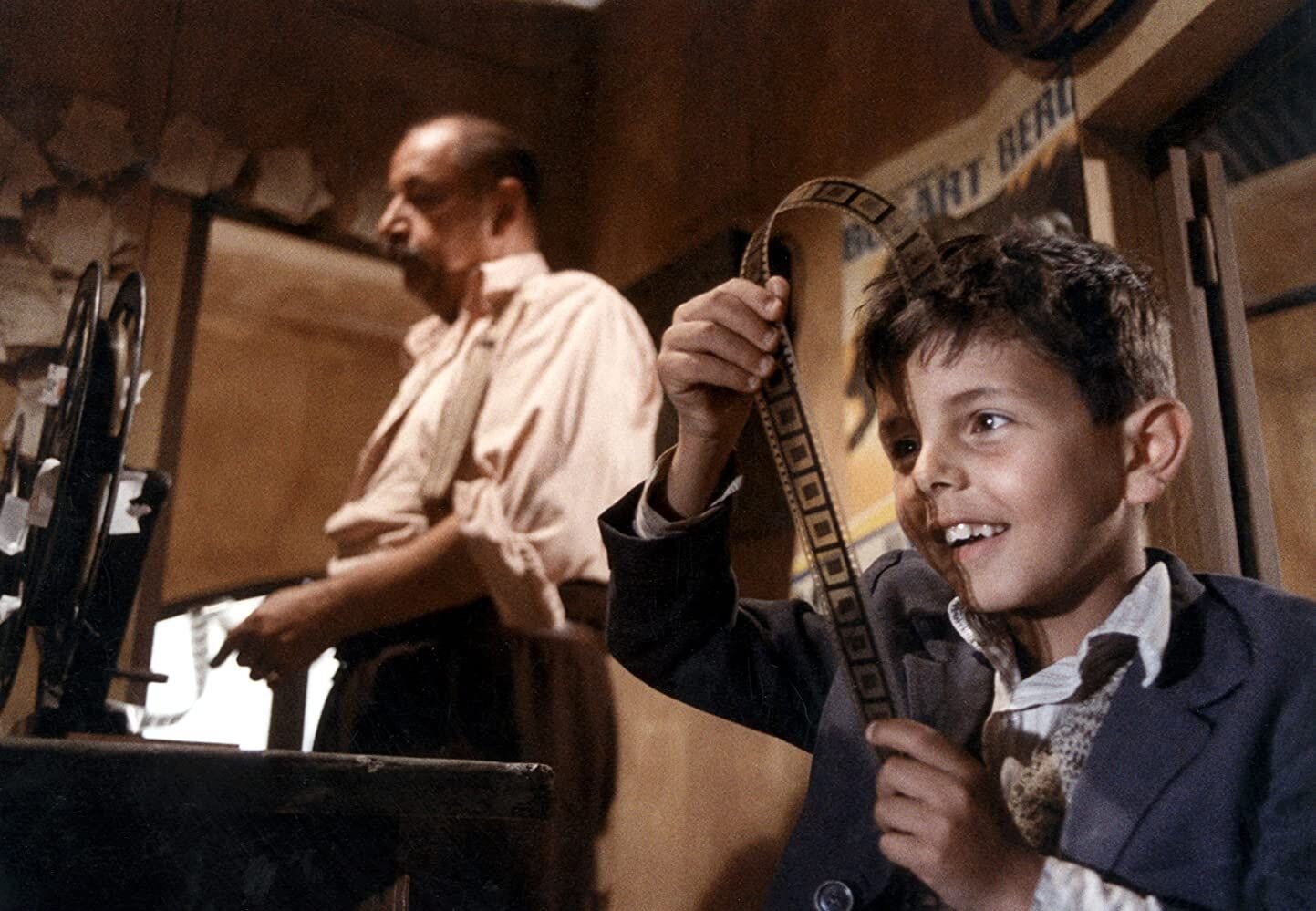 In a still from Cinema Paradiso, we see a boy on the right smiling as he looks at the stills in a roll of film. Behind him, a man works toward projecting a film. In a still from Cinema Paradiso, we see a boy on the right smiling as he looks at the stills in a roll of film. Behind him, a man works toward projecting a film.