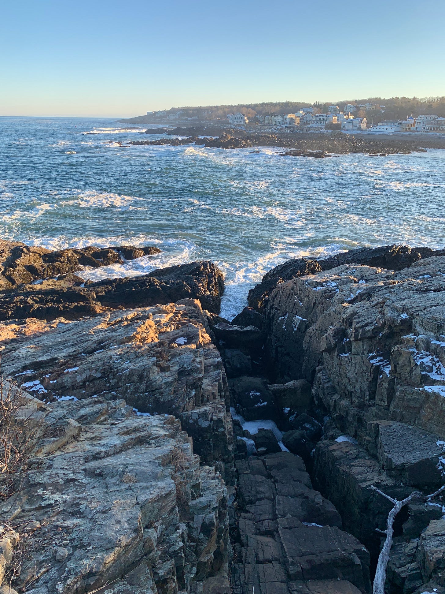 A rocky coast with teal water in late afternoon. In the distance is a piece of coast with houses and buildings on it, in the fore are snowy rocks.