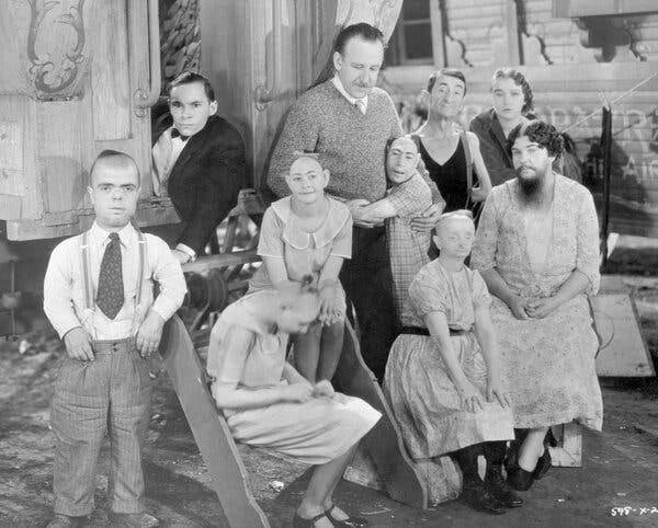 In a black and white photo a man in a shirt, tie and v-neck sweater stands at center, outside a circus trailer, surrounded by actors playing sideshow characters in a 1932 film.