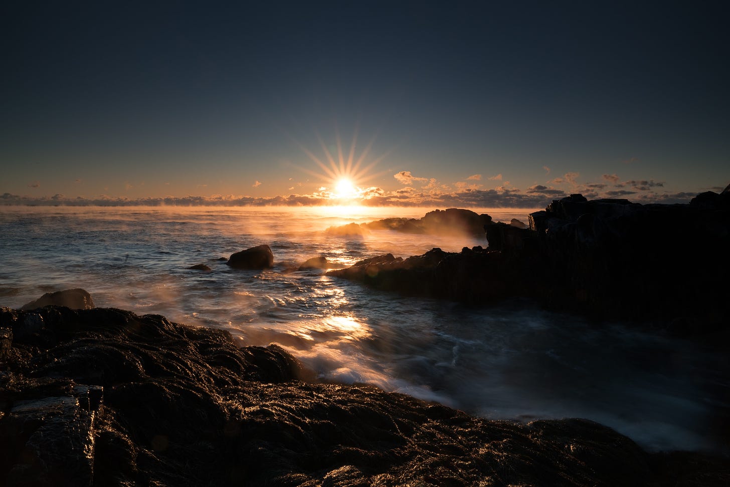 The sun rises in front of gentle waves running up against rocks.