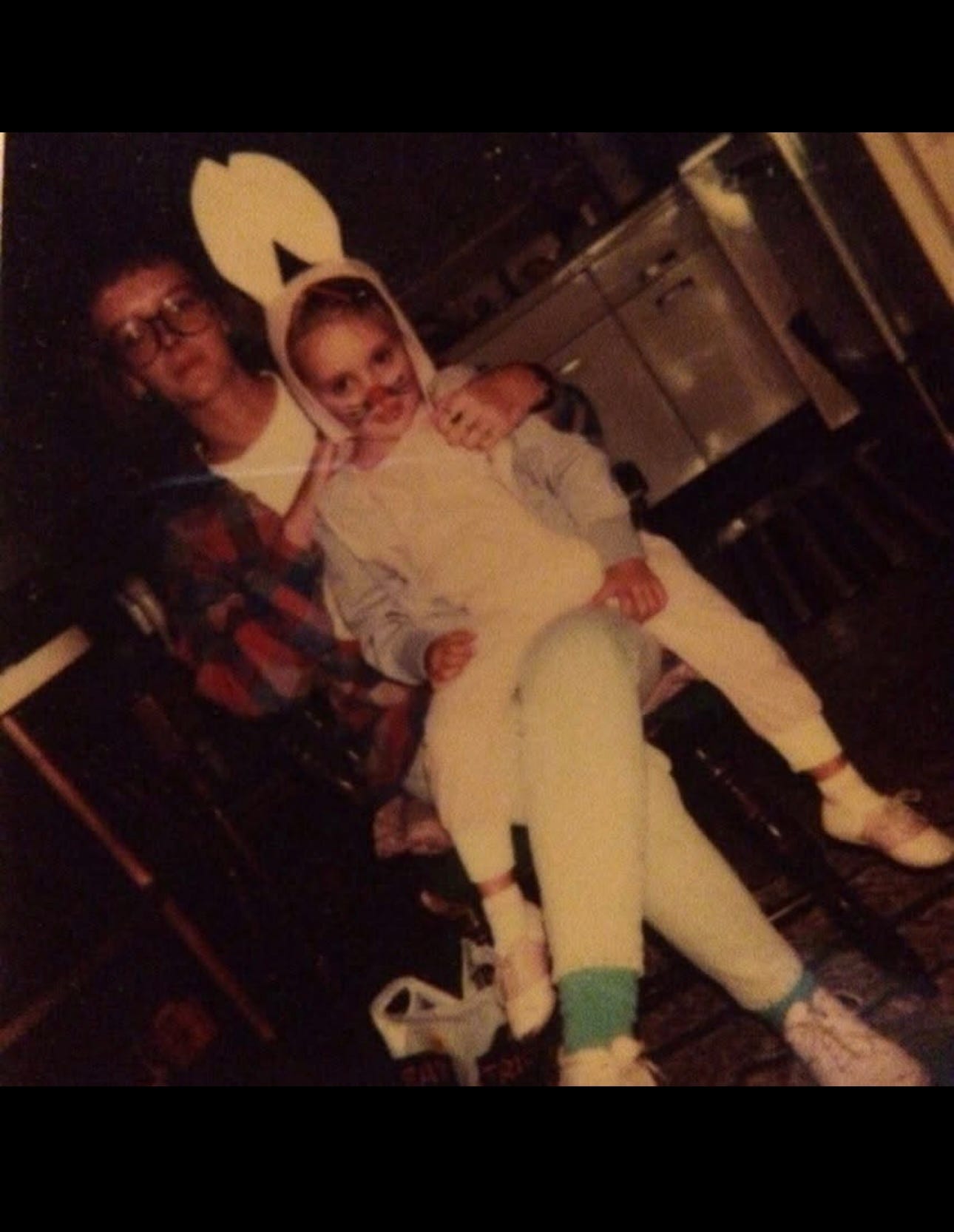 **Alt text:** A vintage photo of a young child dressed in a bunny costume with face paint, sitting on the lap of an adult wearing glasses and a plaid shirt. They are indoors in a warmly lit kitchen, with white cabinets and a dark floor. The moment feels candid and nostalgic, capturing a tender family memory from the past.