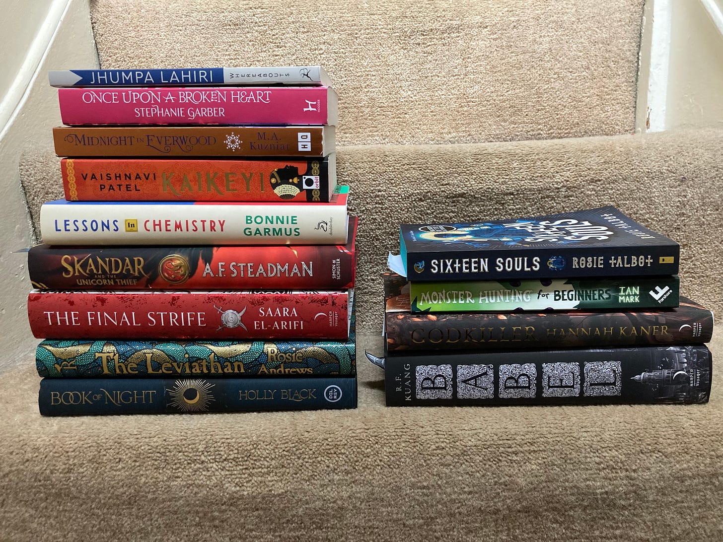 There are two stacks of books situated on carpeted stairs. The one on the left, various titles, is larger than the one on the left, which includes the books mentioned below as read/finished: Babel, Monster Hunters, Godkiller and Sixteen Souls.
