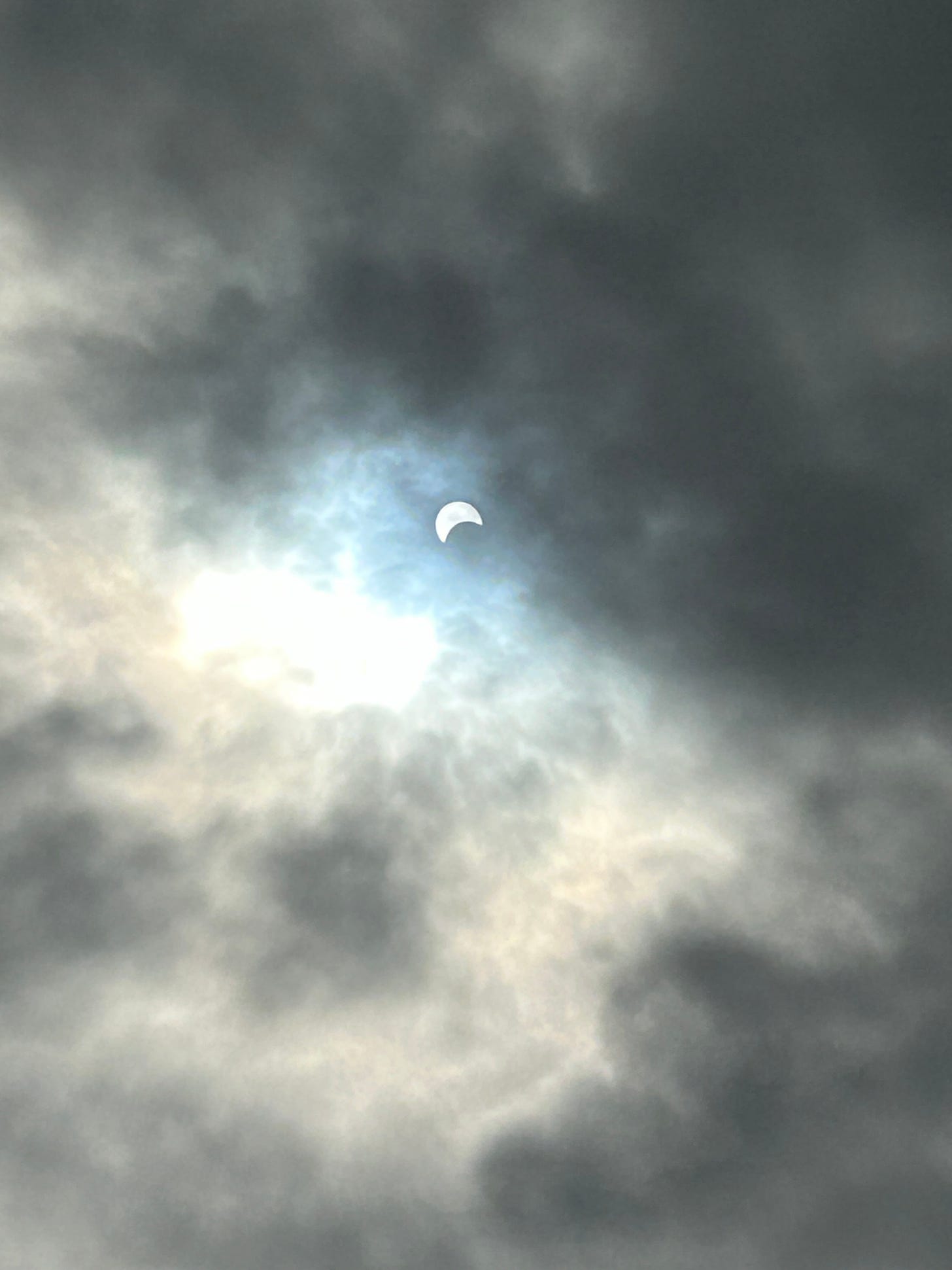 The sun appears as a bright white disk in a cloudy sky with a black crescent—the moon—obscuring the bottom half of it.