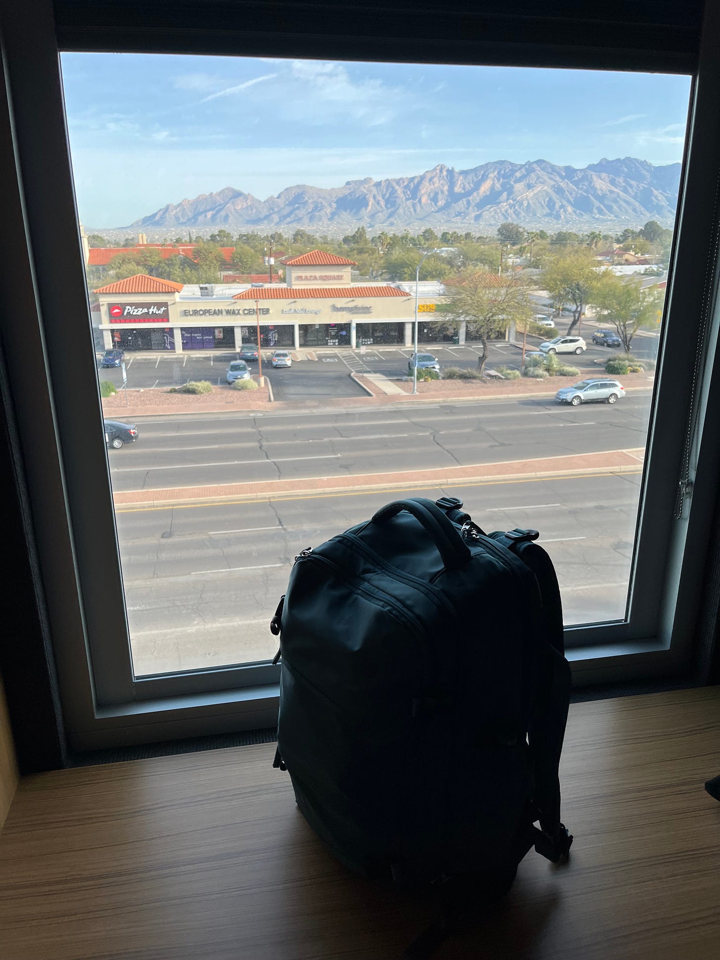Picture of the view outside my hotel window, showing the Catalina mountains, a strip mall, and my single backpack propped up in front of it