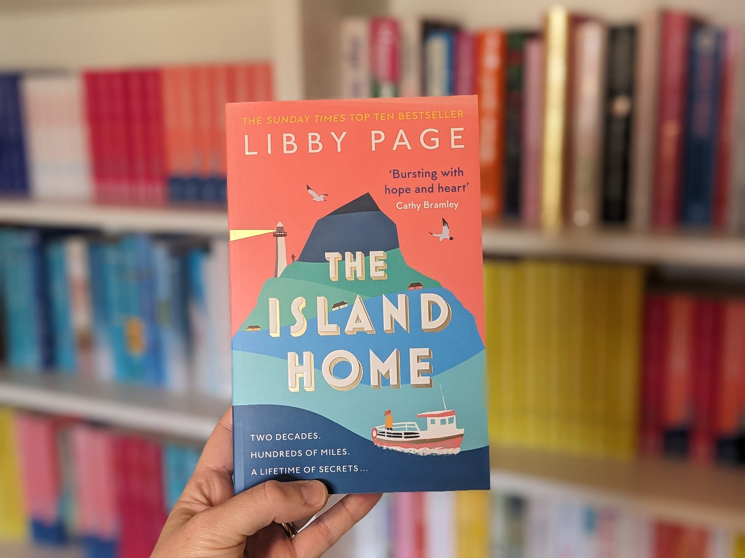 The cover for The Island Home by Libby Page is a salmon pink with an island in the middle in shades of blue. There is a lighthouse and an image of a woman heading towards the island on a boat.