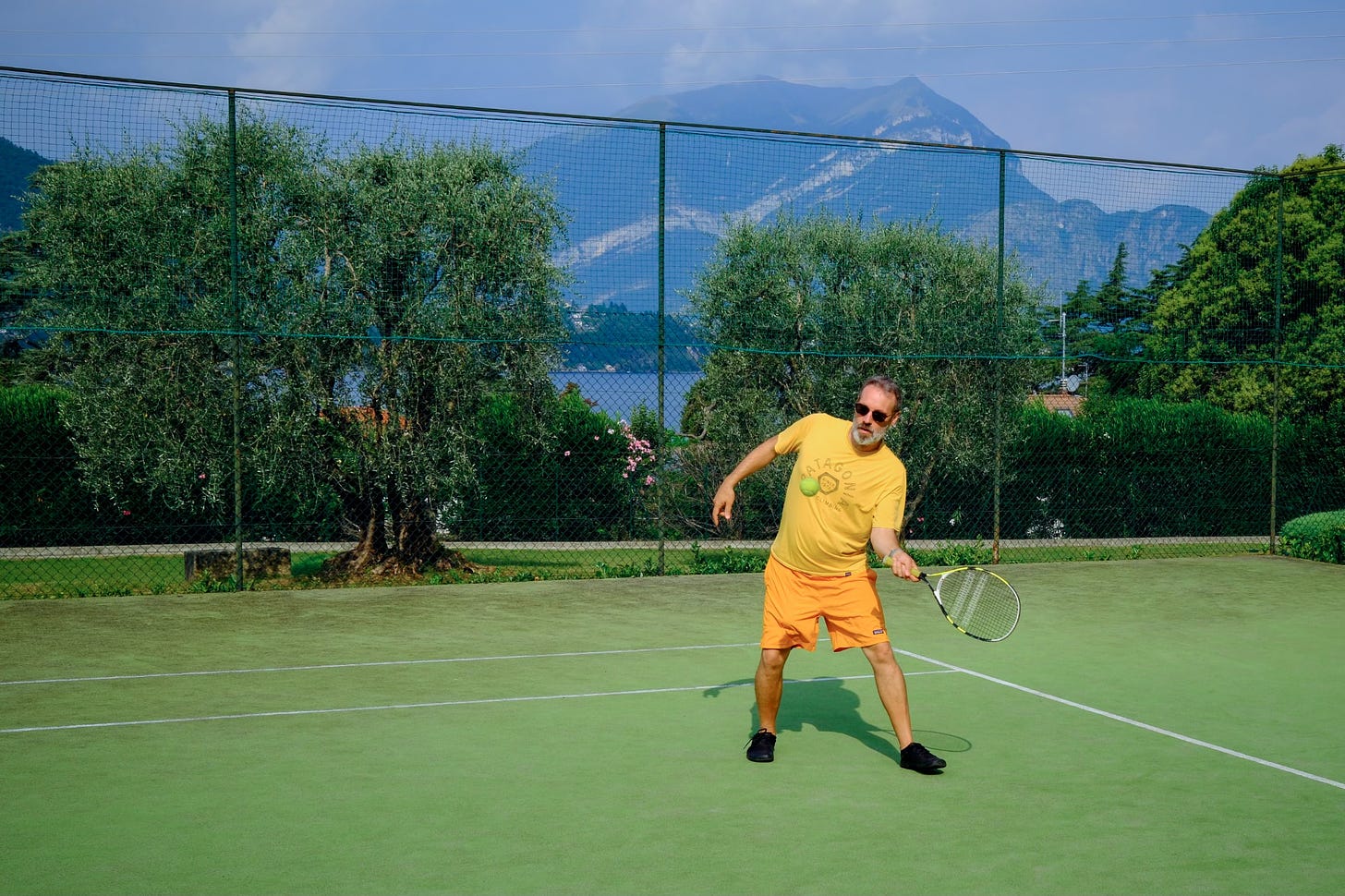 A photo of me playing tennis. It's quite vibrant because it was taken on my X-100IV using the Kodachrome recipe. But if you are visually impaired that might not mean much. I'm wearing a yellow T-shirt, orange shorts and black shoes because I had nothing that matched the otherwise quite summary colours. In the background there is a mountain and although the photograph might look like I know what I'm doing I have not played tennis for over 20 years. That said I did win that particular match ;-) 