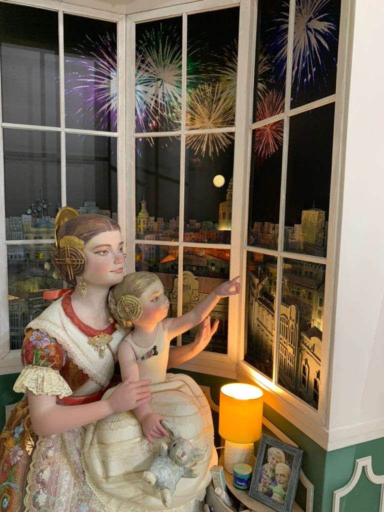 A mother and daughter watch fireworks over the city from their window; the mother is dressed in her full costume, the young daughter has taken the outer dress off, I think in preparation for going to bed after the show