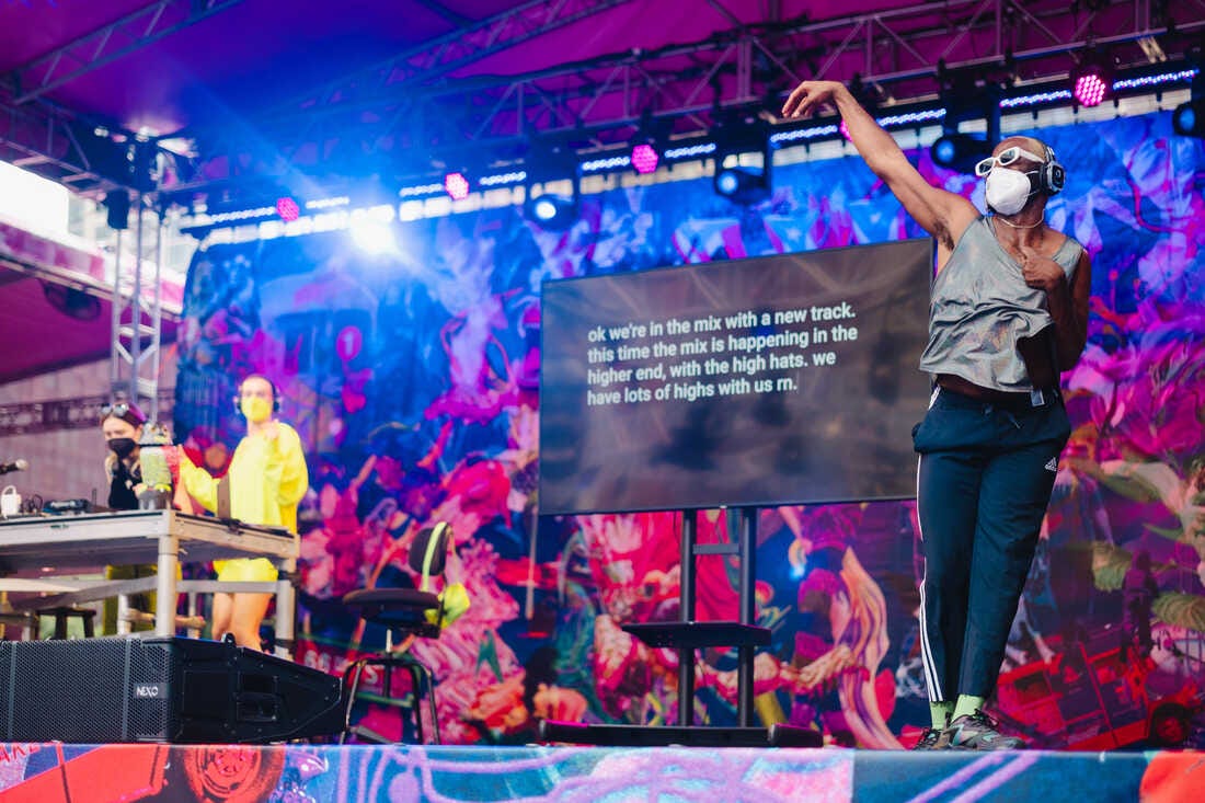 The view of an outdoor stage at Lincoln Center with dancer Jerron Herman, DJs Who Girl and Crip Time, with a screen displaying real-time creative captions, and many colors on the stage's backdrop.