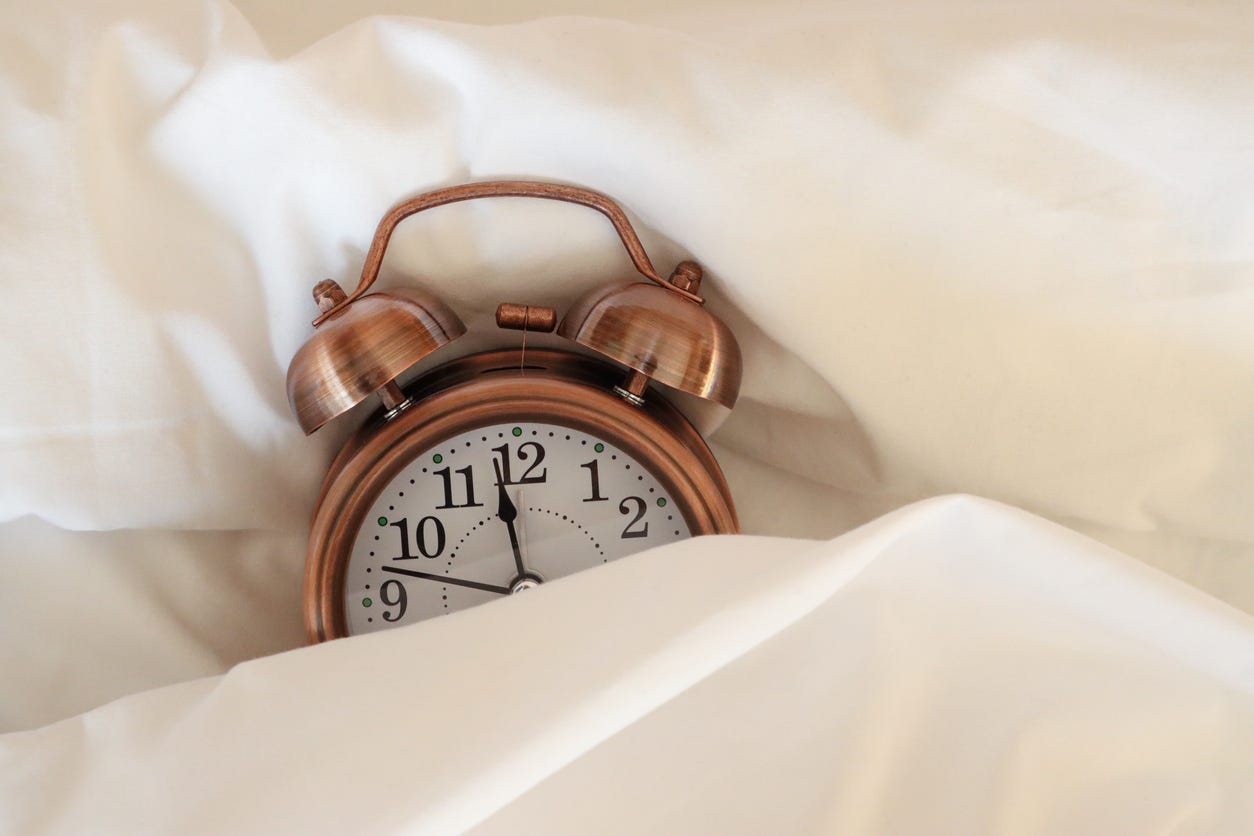Stock photo showing close-up view of retro, double bell, analogue alarm clock tucked up for bedtime on pillow and under bedding.
