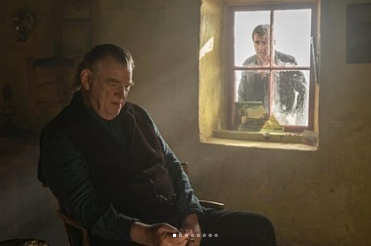 A scene from banshees of Inisherin wirth Brendon Glesson sat on a chair as Colin Farrell looks through the window
