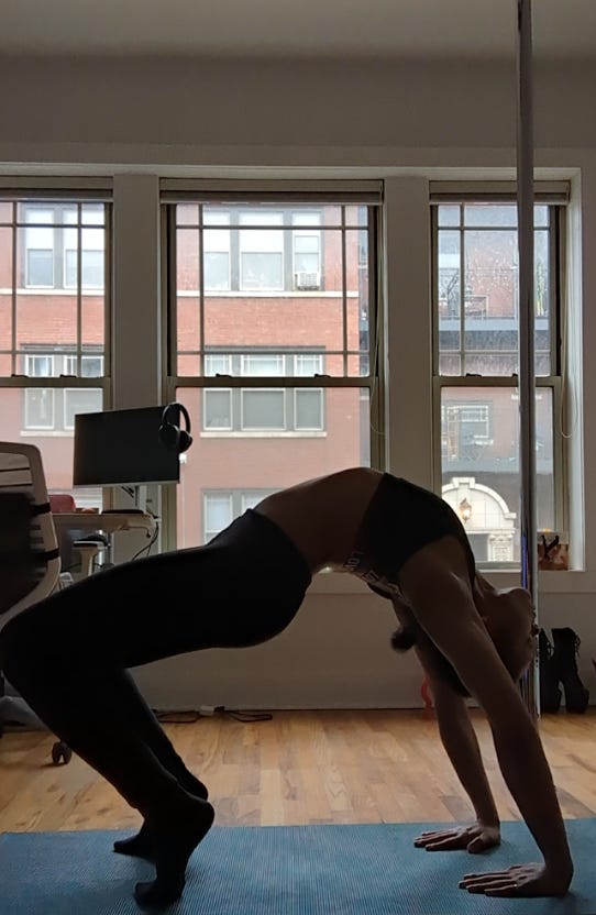 Nathalie attempts a backbend in her living room above her blue yoga mat. She is wearing a black sports bra, black leggings, and black socks. She is in front of big windows making her figure look more shadowy. Her pole and park of her desk is exposed in the background