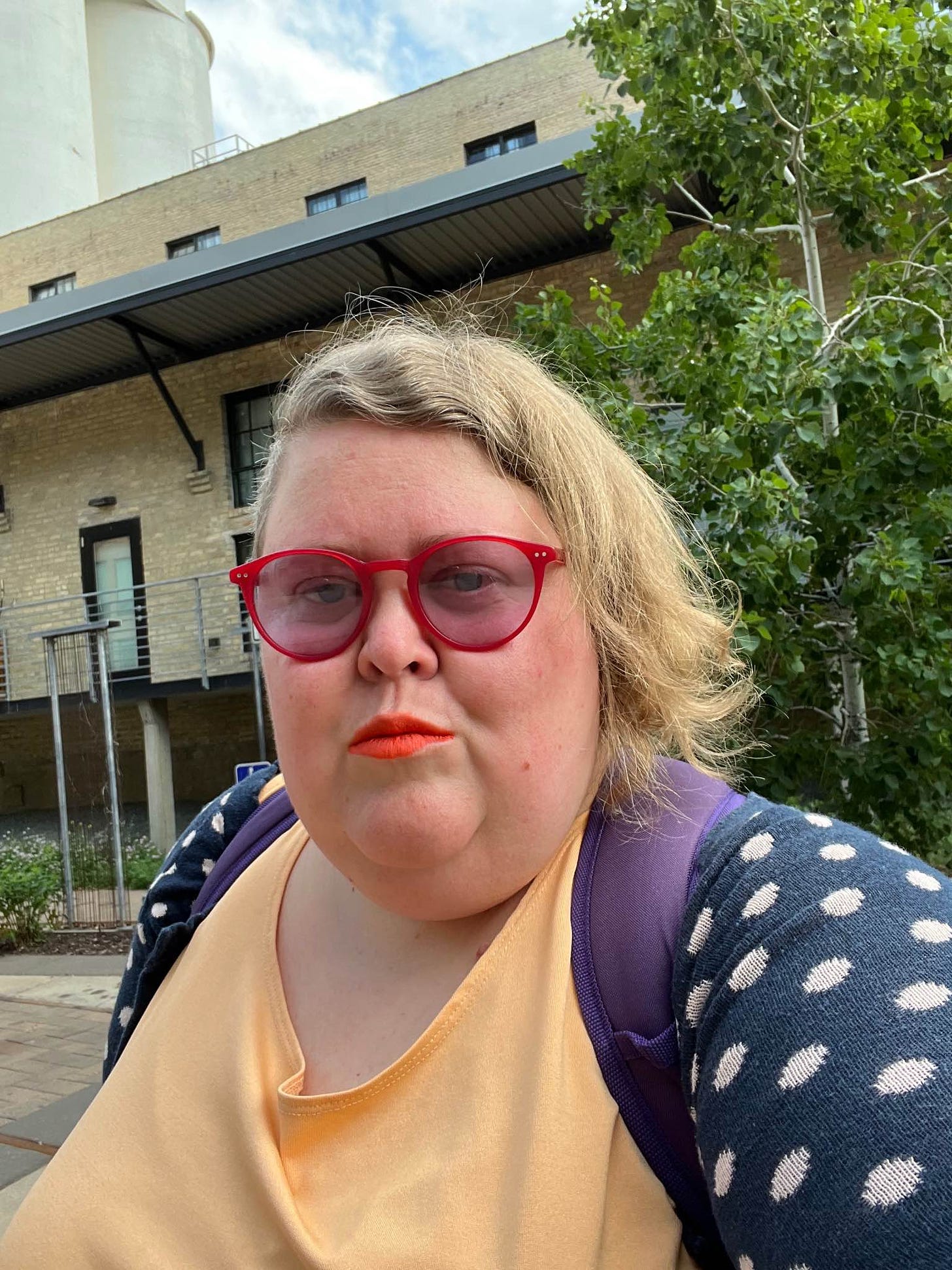 A selfie of me, Alison, a white woman with asymmetrical blonde hair wearing red migraine glasses, a yellow top with a blue polka dot sweater in front of greenery and an old brick building