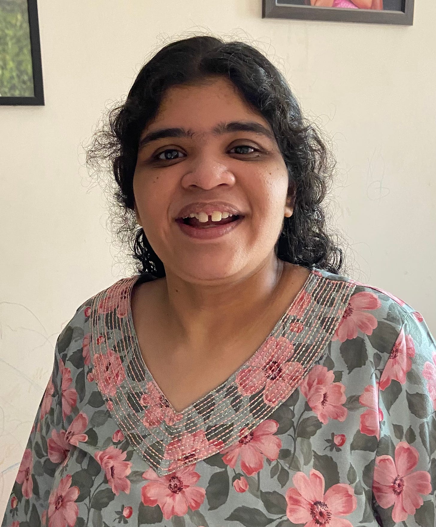 Aditi, a non-speaking autistic Indian teenage girl with long curly black hair is wearing a grey shirt with pink flowers and smiling into the camera. 