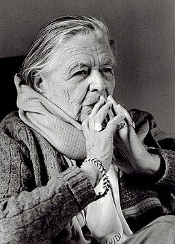 Marguerite Yourcenar, sitting with her hands steepled under her nose, wearing a sweater and a scarf.