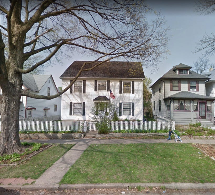 two story white house with a picket fence around the yard and a young child pushing a stroller in front of it at the far end of the right boundary of the yard