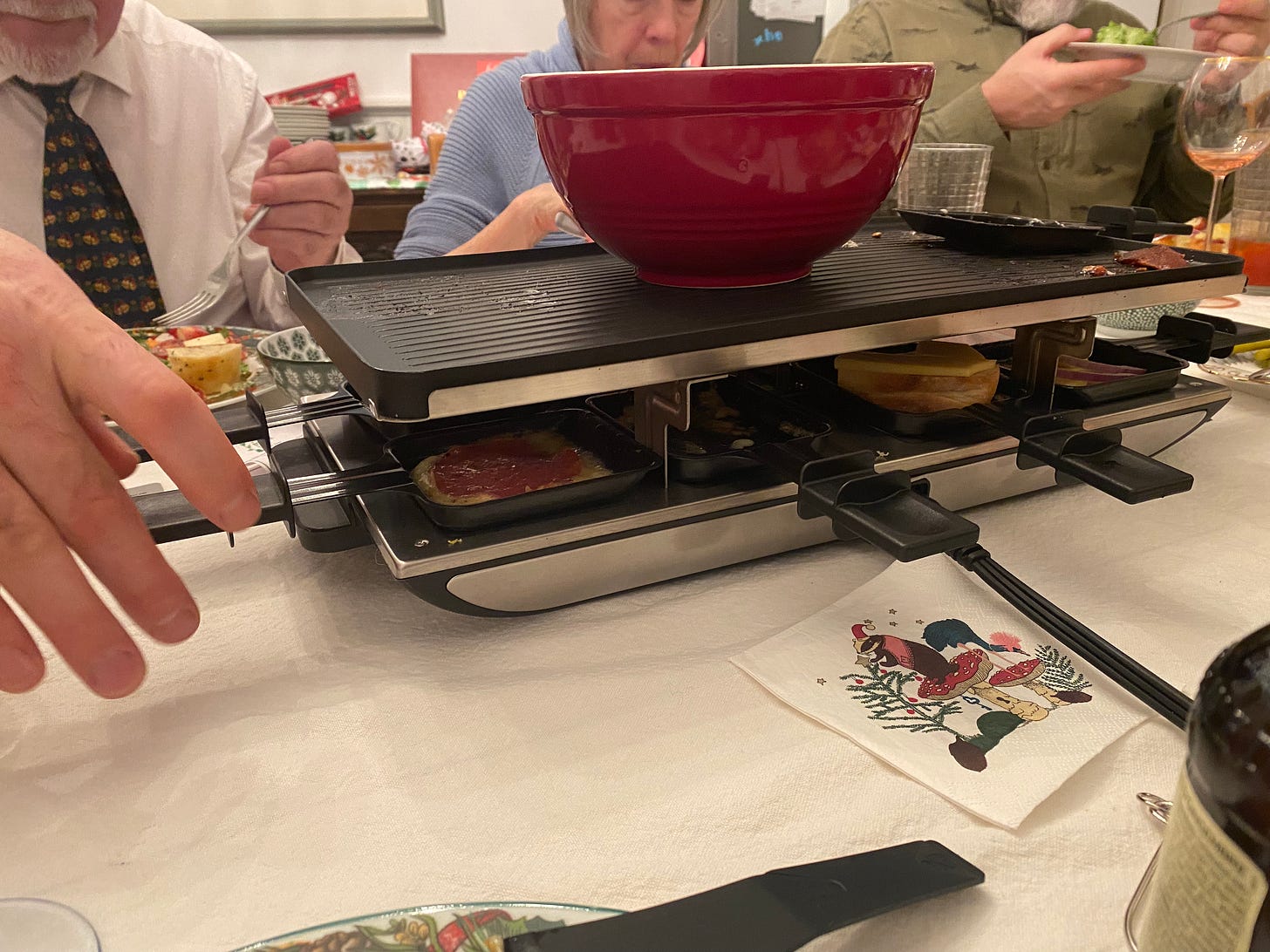 A rectangular raclette set with the paddles in use, a red bowl (of potatoes) warming on top. People are eating on the opposite side of the table. In the foreground a hand reaches toward the set, and a paper napkin with a badger in a Santa hat printed on it sits on the table.