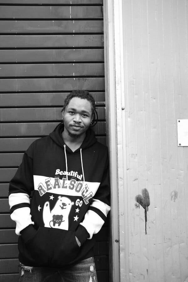 A black and white photograph of the novelist Ani Kayode Somtochukwu, wearing a hooded sweatshirt with an image of two bears on the front, and the words “Beautiful Realsoul Go.” 