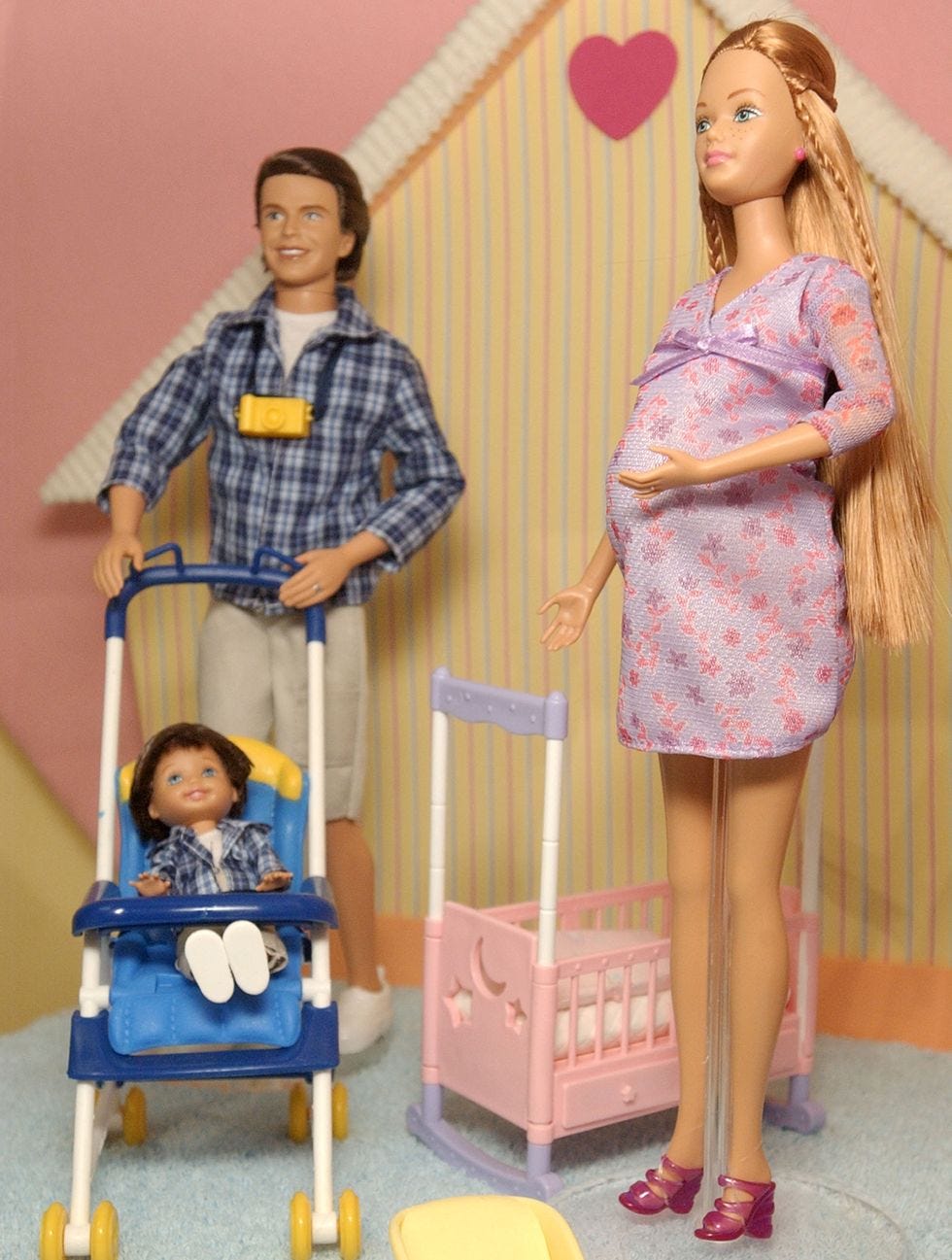 A pregnant Barbie doll stands next to her husband doll who is pushing a child doll in a stroller.
