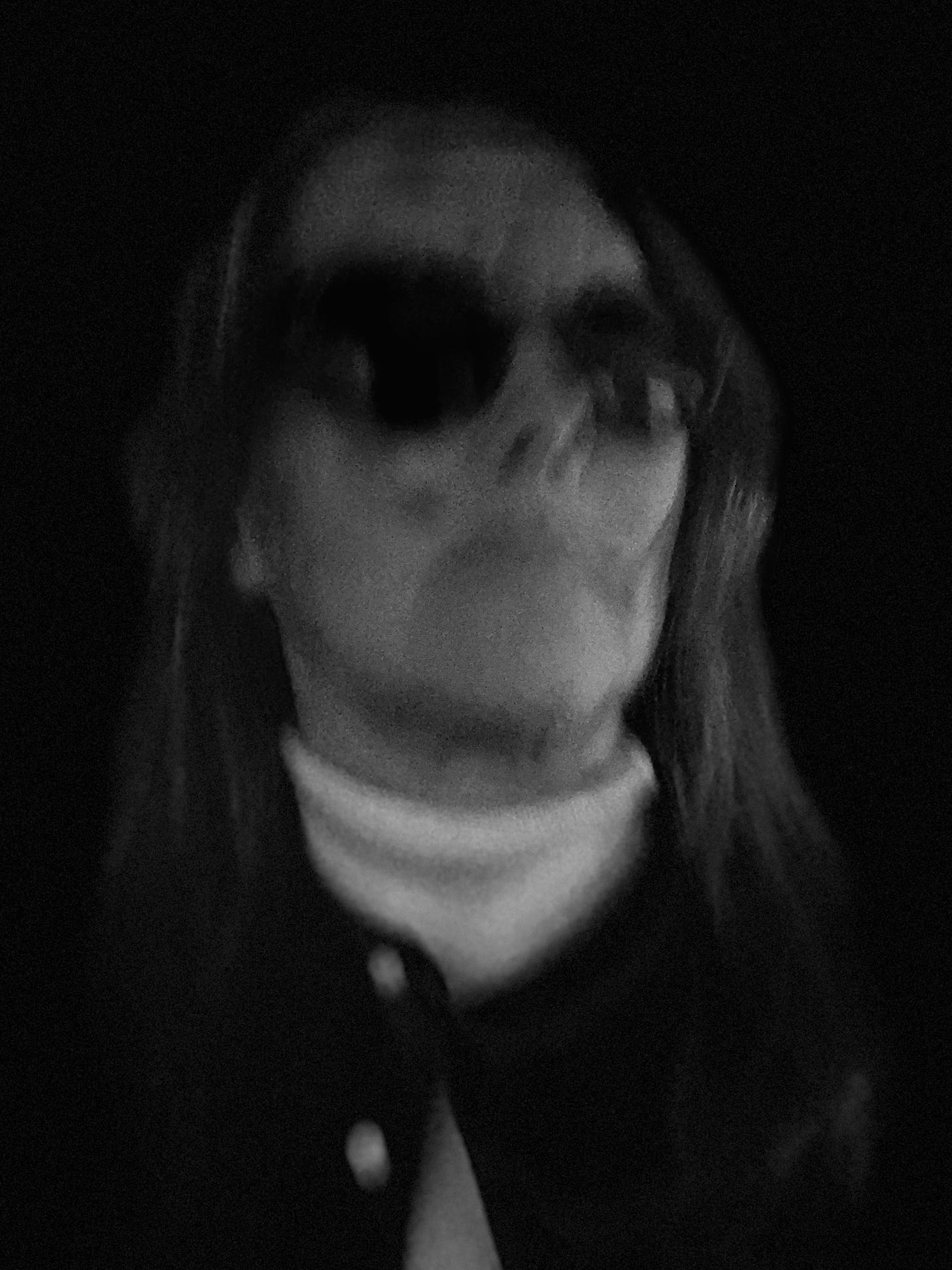 Self portrait of Jody Day. White woman in black shirt and white polo neck and dark glasses. Image is blurred and distorted, and shot in the dark. 